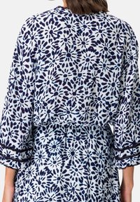 Femme portant une robe blanche avec un motif floral bleu marine et de larges manches avec une bordure en dentelle noire, vue de dos.