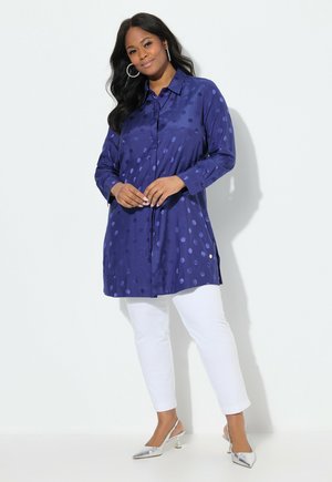 Marineblaues Langarmhemd mit einem Punktenmuster, Knöpfen vorne und Schlitzen an den Seiten, kombiniert mit weißen Hosen und silbernen Absätzen.