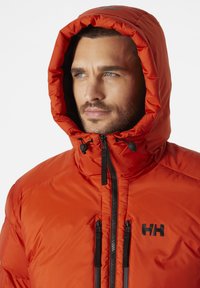 Oranžová izolační bunda s kapucí, se zipem vpředu a černými kovovými detaily; lehký materiál s hladkou texturou.