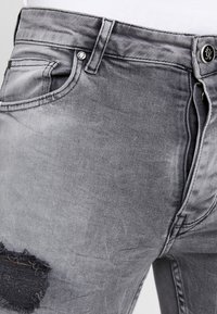 Grå denimjeans med ett slitna mönster, med ett rivet avsnitt. Inkluderar klassisk femficksdesign och metallknappstängning.