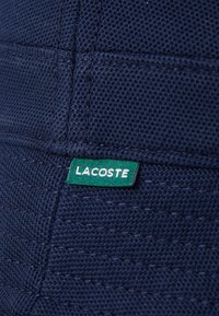 Un tissu texturé bleu marine présente un motif tissé. Une étiquette verte avec le texte blanc "LACOSTE" est cousue sur le côté.