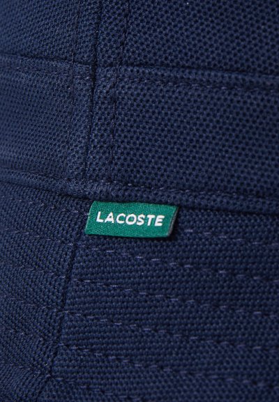 Un tissu texturé bleu marine présente un motif tissé. Une étiquette verte avec le texte blanc "LACOSTE" est cousue sur le côté.