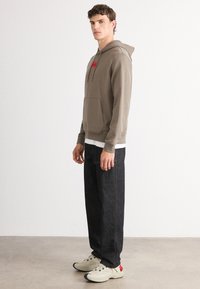 Felpa con cappuccio marrone e tasca frontale, con etichetta rossa. Abbinata a pantaloni di jeans scuri e scarpe beige con dettagli rossi.