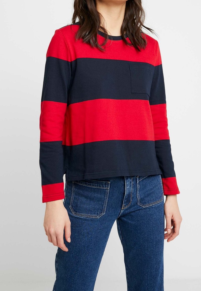 Červeno-modrý pruhovaný dlouhý rukáv se štýlovým kulatým výstřihem a náprsní kapsou, doplněný modrými džíny. Hladká látka, volný střih.