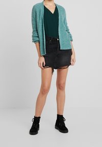 Tealfarbener, flauschiger Cardigan über einem grünen V-Ausschnitt-Top, kombiniert mit einem schwarzen Minirock aus Denim mit fransigen Kanten und schwarzen Schnürstiefeln bis zum Knöchel.