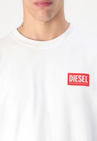 T-shirt en coton blanc avec un col ras du cou, arborant un patch logo rectangulaire rouge sur lequel est inscrit "DIESEL" et "POUR UNE VIE RÉUSSIE."