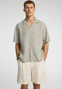 Jasnoszara, krótkiego rękawa koszula lniana z zapięciem na guziki i kołnierzykiem, zestawiona z plisowanymi szortami w kolorze kremowym.