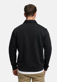 Czarna bluza z kołnierzem, z fakturowanym wykończeniem i prążkowanymi ściągaczami. Tył zdobi okrągły szew. Noszona na białej koszuli.