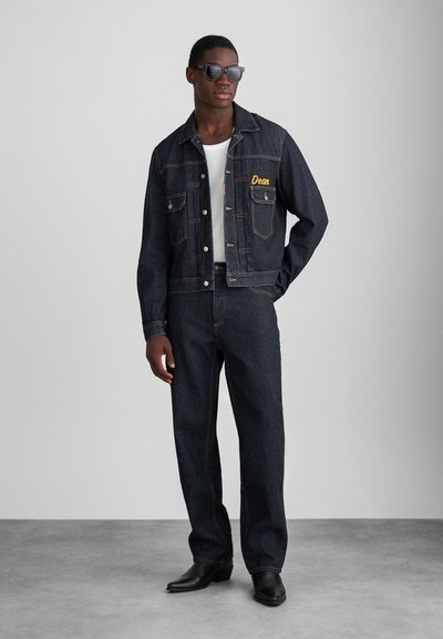 Mørkeblå denimjakke og -bukser med kontrasterende syninger og forlommer. Sorte solbriller og en hvid tanktop fuldender looket.