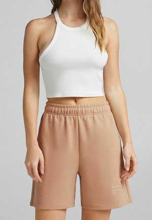 Kvinde iført en hvid ribbet kort top og lysebrune, elastiske, afslappede shorts mod en ensfarvet baggrund.