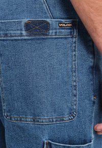 Pantaloni shorts in denim con un lavaggio blu, dotati di una tasca posteriore, cuciture visibili e un'etichetta del marchio Volcom nell'angolo in alto a destra.