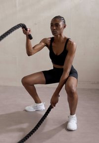 Svart treningsshorts og crop top båret av en person som bruker battle ropes, med hvite treningssko. Innendørs område med en nøytral bakgrunn.