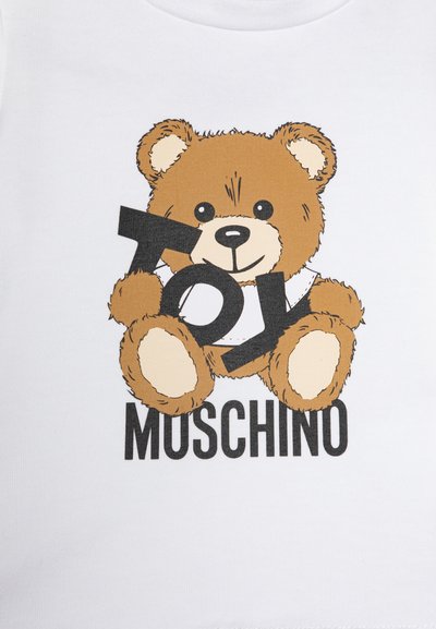 Valkoinen paita, jossa on ruskean karhun graafinen kuva, joka pitää mustilla paksuilla kirjaimilla tekstiä "TOY", sen alla on musta teksti "MOSCHINO".