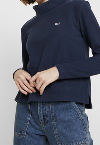 Maglia a maniche lunghe blu navy con colletto alto, caratterizzata da un piccolo logo sul petto. Il tessuto appare morbido con spacco laterale all'orlo.