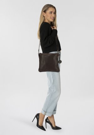 Borsa a tracolla in pelle marrone scuro con design piatto, accenti in metallo e un charm laterale. Il modello la indossa con jeans chiari e tacchi neri.