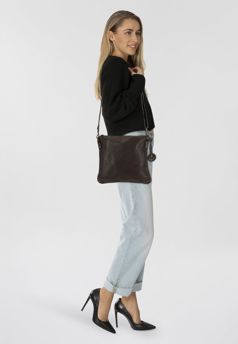Borsa a tracolla in pelle marrone scuro con design piatto, accenti in metallo e un charm laterale. Il modello la indossa con jeans chiari e tacchi neri.