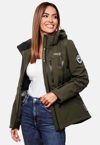 Woman smiling, wearing an olive green waterproof jacket over a white shirt and blue jeans, with jacket partially unzipped.