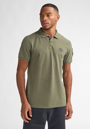 Homme portant un polo vert olive avec un logo sur la poitrine et un pantalon noir, debout devant un fond blanc uni, regardant sur le côté.