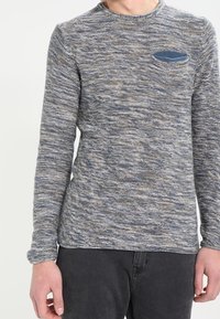 Chemise en tricot à manches longues dans des tons de bleu et de gris, avec un motif texturé et une petite poche poitrine avec un accent bleu.