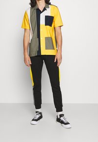 Kortärmad, flerfärgad skjorta med en blandning av gula, gröna, marinblå och beige rutor; ihop med svarta byxor med gula sidan.