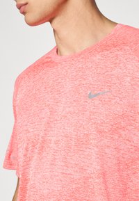 Hombre con camiseta deportiva Nike de color coral jaspeado, con pequeño logo de swoosh gris en el lado izquierdo del pecho, visible desde el cuello hasta la mitad del torso.