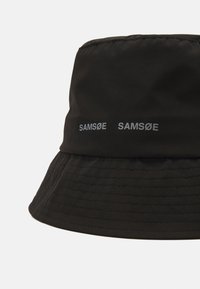 Svart bøttehatt laget av glatt stoff, med en sammenleggbar skjerm, teksturert overflate og to hvite "SAMSØE"-logoer trykket på siden.