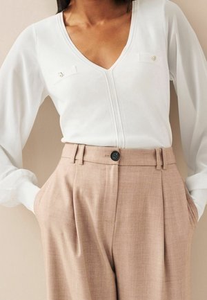 Femme portant un haut blanc à manches longues avec un col en V et des poches décoratives sur la poitrine, ainsi qu'un pantalon plissé taille haute beige avec un bouton noir.