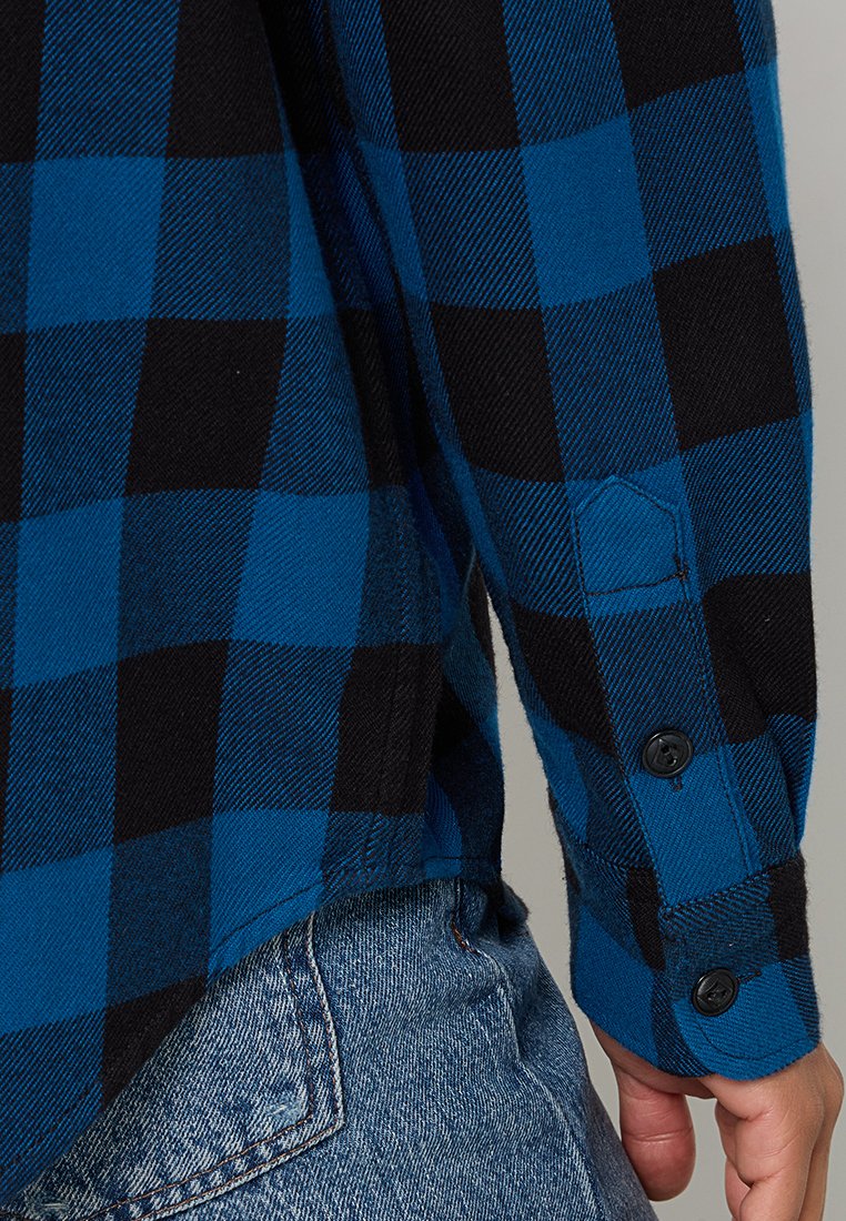 Camicia di flanella a scacchi blu e nera con polsini abbottonati, caratterizzata da un tessuto testurizzato e un orlo arrotondato. Jeans in denim parzialmente visibili.