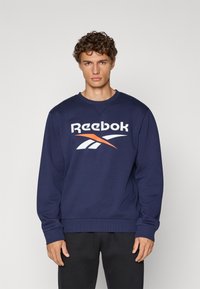 Námornícky modrý sveter Reebok s okrúhlym výstrihom, dlhými rukávmi a veľkým bielym a oranžovým logom cez hruď. Hladká textúra látky.