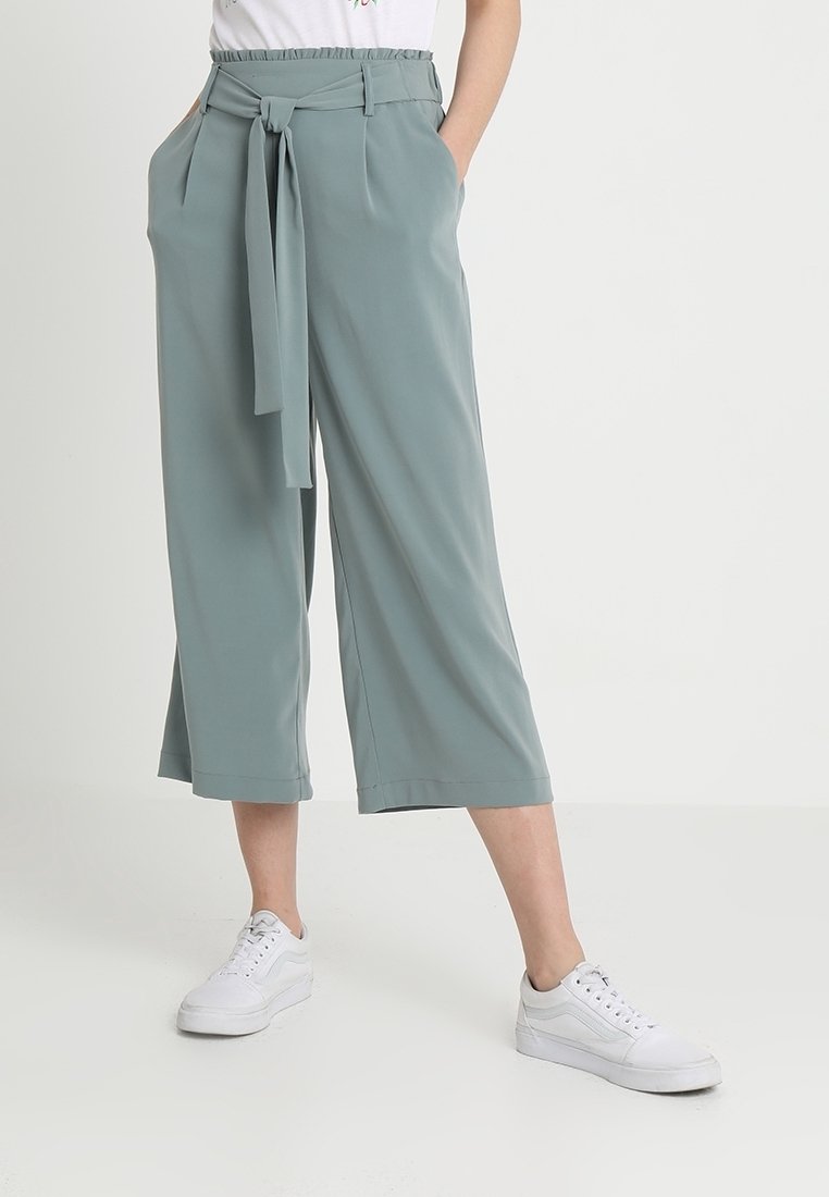 Femme portant un pantalon large, court, vert clair avec une ceinture nouée à la taille et des baskets blanches, debout, les mains dans les poches.