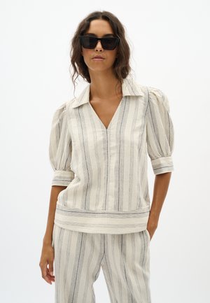 Vrouw draagt creme gestreepte blouse met pofmouwen en bijpassende broek, gecompleteerd met grote zwarte zonnebril.