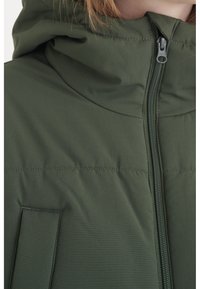 Grön vadderad jacka med hög krage, dragkedjestängning och sydda detaljer på tyget. Har en framficka på vänster sida.