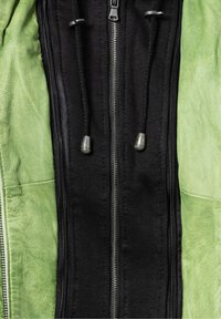 Grüne Lederjacke mit einem schwarzen Baumwollmittelteil, ausgestattet mit einem Reißverschluss und Kordelzügen am Kragen, die kontrastierende Texturen zeigen.