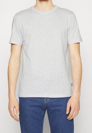 Mann trägt ein hellgraues kurzärmeliges T-Shirt mit kleinem "BOSS"-Logo auf der Brust und blaue Jeans, steht mit entspannten Armen.