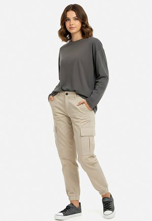 Młoda kobieta stoi z rękami w kieszeniach, ubrana w ciemnoszarą bluzkę z długim rękawem, beżowe spodnie typu cargo i czarne trampki na białym tle.