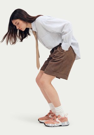 Young woman bending forward wearing a striped shirt, brown shorts, white socks, and pink sneakers with thick soles.
