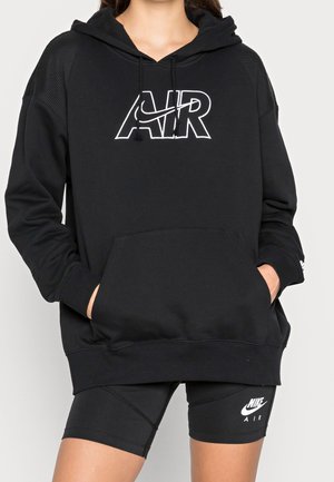 Mujer que lleva un hoodie negro de Nike Air con bolsillo canguro y pantalones cortos deportivos negros de Nike Air, con las manos en los bolsillos del hoodie, cortado a la altura de los hombros y los muslos.