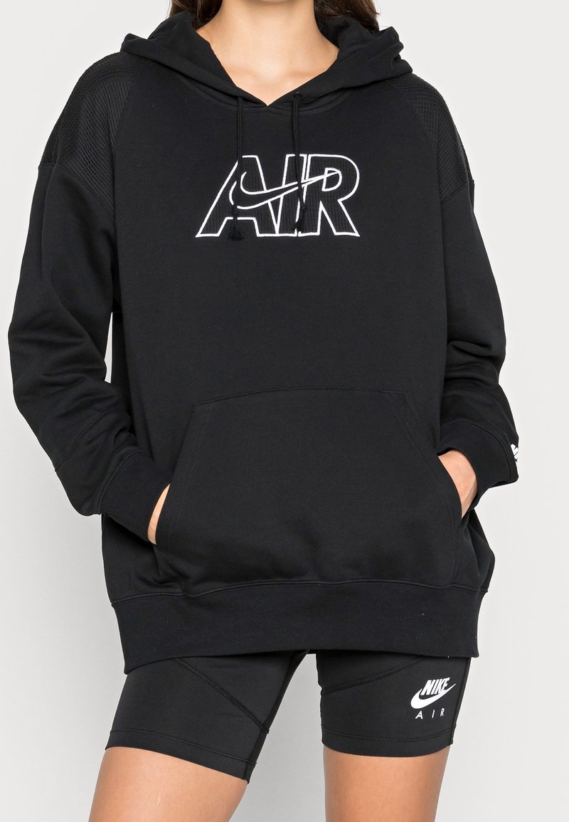 Kvinna som bär en svart Nike Air-hoodie med kengurufickor och svarta Nike Air-träningsshorts, händerna i hoodiefickorna, klippt vid axlarna och låren.
