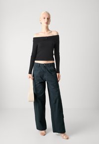 Svart off-shoulder topp med lange ermer, kombinert med mørkegrønne vide bukser. Modellen holder en beige veske og står mot en lys bakgrunn.