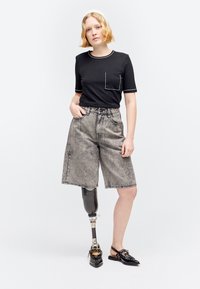 Jeune femme debout avec une jambe gauche prothétique, portant une chemise noire avec une poche, un short gris mi-genou et des chaussures noires sans lacets.