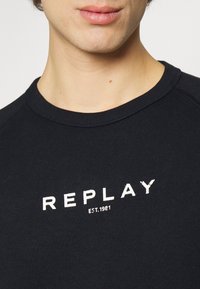 T-shirt nera in cotone con collo tondo, con logo "REPLAY" bianco stampato e la scritta "EST. 1981" sottostante, texture liscia, vista ravvicinata.