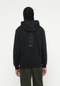 Braidelt hajjal rendelkező személy fekete Nike kapucnis pulóverben, amelyen függőleges logó és swoosh található a hátsó részen, sötétzöld cargo nadrággal párosítva.
