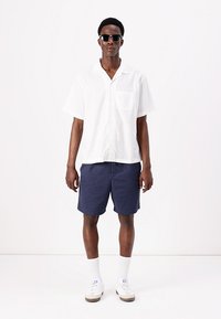 Hvit kortermet skjorte med knapper og lomme, kombinert med mørk marineblå shorts og hvite joggesko. Modellen har på seg svarte solbriller.