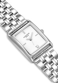 Hõbedane ristkülikukujuline kell tekstureeritud valge näo ja hõbedaste tunni näitajatega. Omab metallist link-rannet ja krooni küljes.