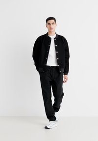 Veste bomber noire avec des boutons-pression blancs, portée sur un t-shirt blanc, associée à un pantalon noir et des baskets blanches. Design minimaliste.