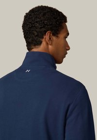 Polo de color azul marino con cuello alto, que presenta una tela texturizada y un pequeño logo blanco en la parte trasera cerca del escote.