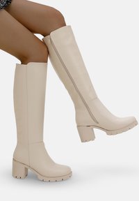 Botas beige hasta la rodilla con un acabado de cuero suave, tacón cuadrado y cremallera lateral; con una suela resistente y con un dibujo pronunciado.