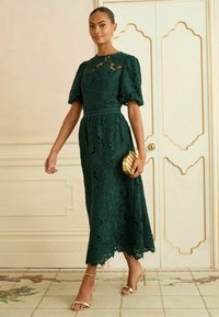 Zaļš mežģīņu midi kleita ar īsām pūkainām piedurknēm, ziedu rakstiem un viļņota apakšmalu. Pievienots zelta rokassomu un siksnu papēžu kurpes.