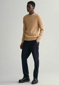 Beiger Kabelstrickpullover mit gerippten Bündchen und Saum, kombiniert mit navyfarbenen Hosen und schwarzen, klobigen Stiefeln. Einfaches, klassisches Design.