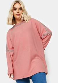 Ylivoimainen vaaleanpunainen huppari, jossa on pyöreä pääntie, hopeanväriset niittikoristeet harteilla ja hihoissa, pehmeää materiaalia.